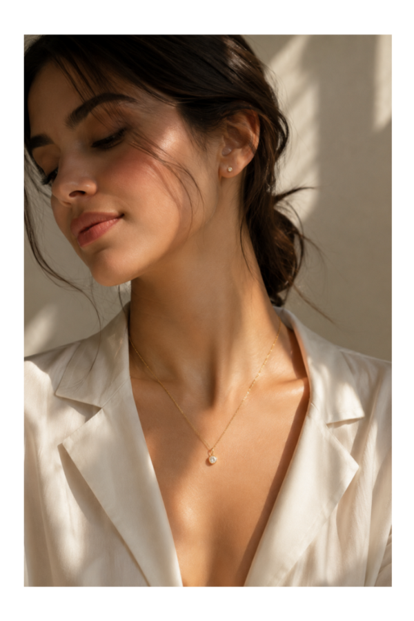 A woman wearing a delicate gold necklace with a small pearl pendant, styled with a white satin blazer under warm, sunlit shadows.